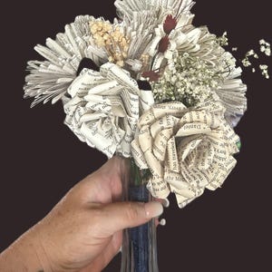 May include: A bouquet of handcrafted flowers made from vintage book pages, featuring folded paper roses and fan-shaped blooms. The arrangement is accented with dried white baby's breath and small dark red sprigs, all tied with a dark blue ribbon.