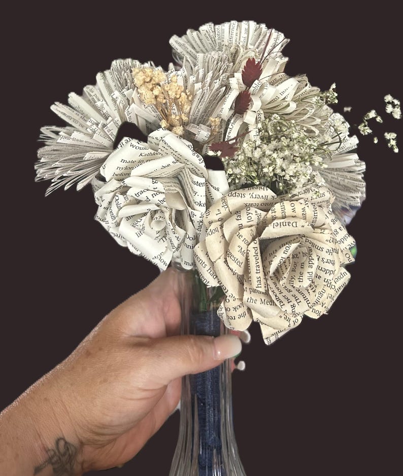 May include: A bouquet of handcrafted flowers made from vintage book pages, featuring folded paper roses and fan-shaped blooms. The arrangement is accented with dried white baby's breath and small dark red sprigs, all tied with a dark blue ribbon.