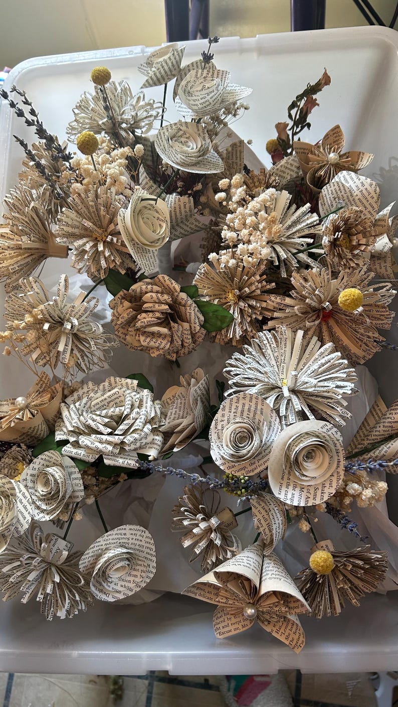 May include: A decorative bouquet of handcrafted flowers made from vintage book pages, featuring various rose and daisy shapes. The arrangement includes dried lavender sprigs, small white baby's breath, and yellow billy ball accents.