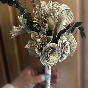 May include: A decorative bouquet crafted from vintage book pages, featuring folded paper flowers, dried lavender sprigs, and dark green eucalyptus leaves. The stems are wrapped in white lace trim, creating a textured arrangement with a neutral color palette.