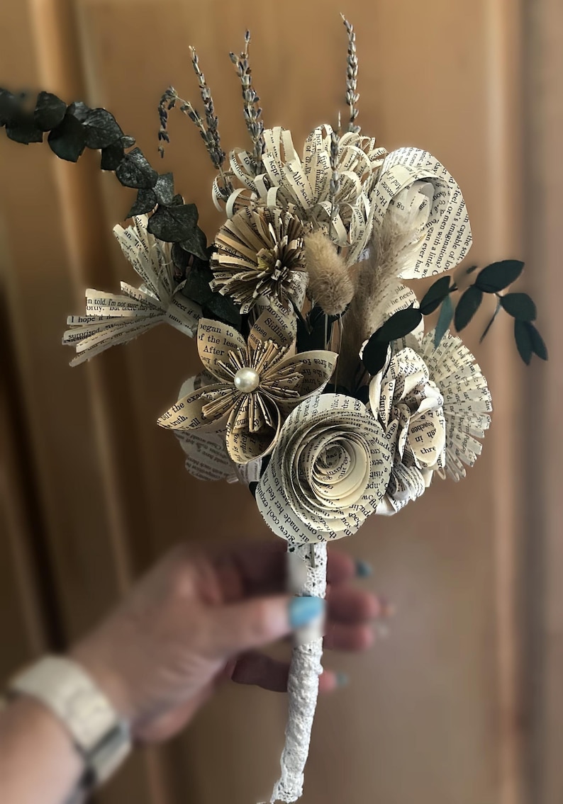 May include: A decorative bouquet crafted from vintage book pages, featuring folded paper flowers, dried lavender sprigs, and dark green eucalyptus leaves. The stems are wrapped in white lace trim, creating a textured arrangement with a neutral color palette.
