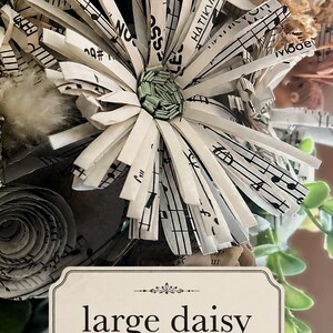 May include: A handcrafted daisy flower made from strips of white paper printed with black musical notation. The center of the flower features small, light green cylindrical elements. A rectangular label below reads large daisy.