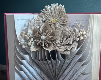 May include: A book sculpture featuring folded pages forming a fan-like base, topped with three paper flowers crafted from book pages. The flowers are accented with small white dried flowers, a central pearl bead, and a small yellow center detail.