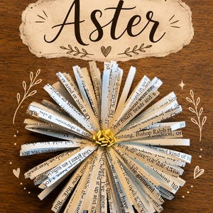 May include: A decorative paper flower crafted from strips of printed book pages with black text, featuring a small yellow center. Above the flower, a beige label with a torn-edge design displays the word Aster in black script with a small heart.