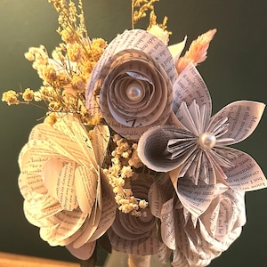 May include: A bouquet of handcrafted paper flowers made from printed book pages, featuring rolled petals and central white pearl accents. The arrangement is interspersed with sprigs of dried yellow baby's breath and light pink grasses.
