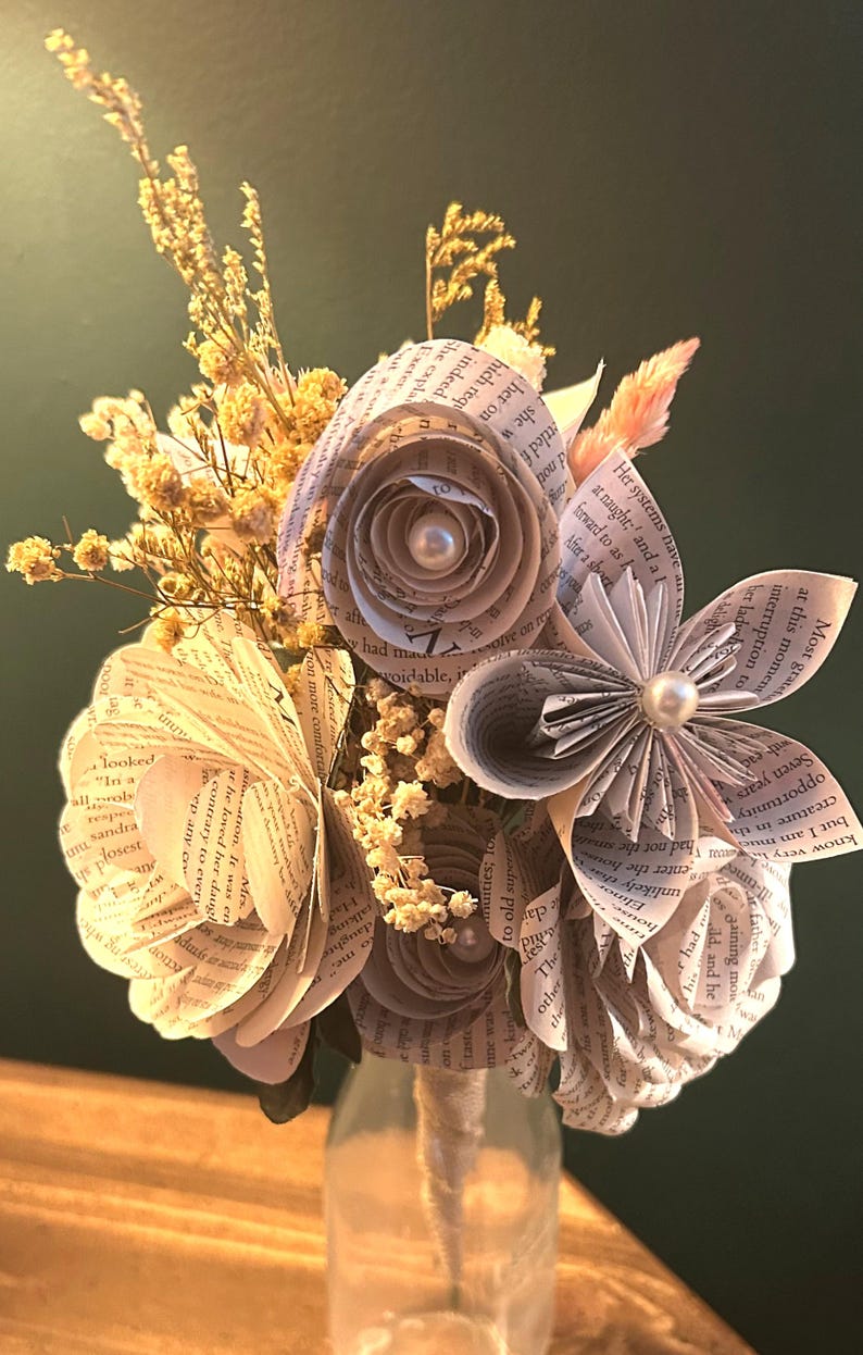 May include: A bouquet of handcrafted paper flowers made from printed book pages, featuring rolled petals and central white pearl accents. The arrangement is interspersed with sprigs of dried yellow baby's breath and light pink grasses.