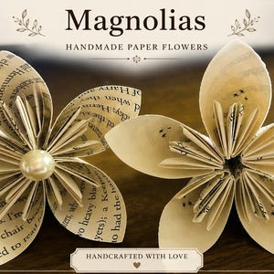 May include: Two handmade paper flowers crafted from book pages with printed text. The left flower features a central cream-colored pearl bead. The text above reads Magnolias Handmade Paper Flowers and the bottom text reads Handcrafted With Love.