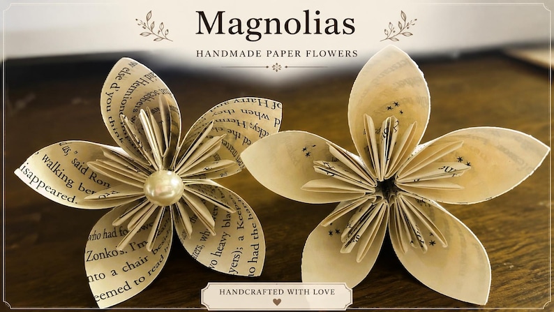 May include: Two handmade paper flowers crafted from book pages with printed text. The left flower features a central cream-colored pearl bead. The text above reads Magnolias Handmade Paper Flowers and the bottom text reads Handcrafted With Love.