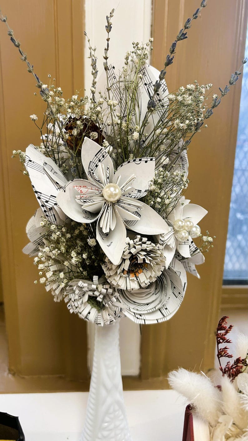 May include: A bouquet of handcrafted paper flowers made from vintage sheet music, featuring white petals with black musical notation. The arrangement includes dried lavender, baby's breath, and central pearl accents, all displayed in a white vase.