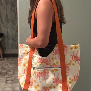 May include: A white tote bag featuring a pattern of pink unicorns and yellow flowers. It has orange straps and a zipped pocket. The bag is being held by a person.