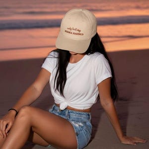 May include: A beige baseball cap with the text "only ball my MAMA RAISED" in brown script. The cap is worn by a person wearing a white t-shirt and denim shorts. The background is a beach at sunset.