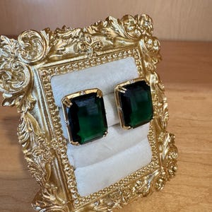 May include: A pair of rectangular emerald green gemstone earrings set in gold metal prongs, displayed on a white velvet ring holder inside an ornate, rectangular gold-colored frame with intricate scrollwork and decorative relief detailing.