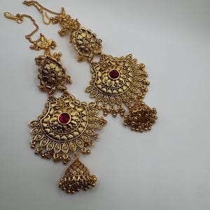 Peut inclure: Une paire d'accessoires de cheveux en métal doré avec des motifs en filigrane complexes, une pierre rouge circulaire centrale et une breloque en forme de cloche suspendue à la base. Chaque pièce inclut une fine chaîne dorée avec un crochet.