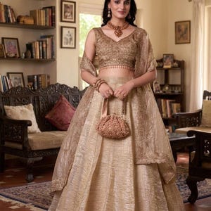 Puede incluir: Un conjunto de lehenga choli dorado que incluye una blusa sin mangas con bordados intrincados y una falda larga a juego con acabado texturizado. El conjunto incluye un dupatta transparente con bordes bordados y un pequeño bolso de cuentas.