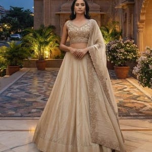 Pode incluir: Um conjunto lehenga choli bege composto por uma blusa sem mangas com bordados dourados intrincados e uma saia longa a condizer. Uma dupatta coordenada com uma borda larga e ricamente bordada está drapeada sobre um ombro.