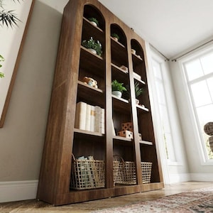 May include: A tall, dark brown wooden bookcase featuring three vertical sections with arched tops. The unit contains multiple open shelves holding various items, including books, small potted green plants, a teddy bear, and woven baskets.