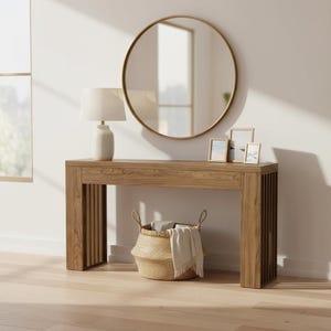 Puede incluir: Una mesa consola rectangular de madera con paneles laterales de listones, coronada con una lámpara de mesa blanca y tres marcos de fotos. Un espejo redondo con marco dorado cuelga sobre la mesa y una cesta tejida con tela blanca está en el suelo.