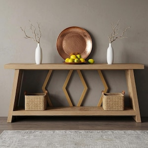 May include: A light oak console table with a geometric gold metal base, featuring two white vases with bare branches, a large hammered copper platter, a bowl of yellow lemons, and two woven baskets on the lower shelf for storage.
