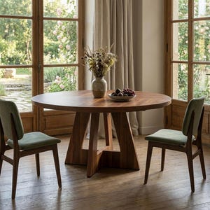 Può includere: Un tavolo da pranzo rotondo in legno con base centrale, accompagnato da due sedie con struttura in legno scuro e sedute imbottite color verde salvia. Un vaso in ceramica con fiori secchi e una ciotola di uva poggiano sul piano.