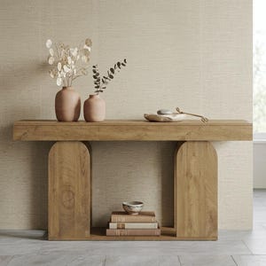May include: A light brown wooden console table with two arched supports. On top are two matte peach vases with dried stems and a stone bowl with a metal tool. A small bowl sits on a stack of three books on the lower shelf of the wooden table.