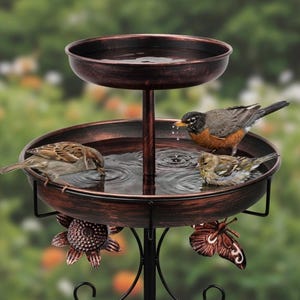 May include: A two-tiered bird bath with a dark bronze finish and copper-toned highlights. The structure features two round basins for water, decorated with metal sunflower and butterfly accents, supporting three small birds drinking water.