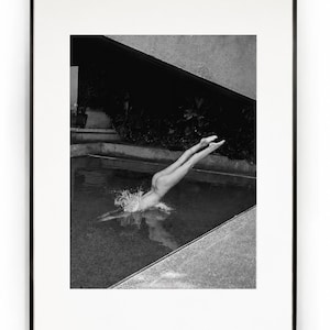 May include: A black and white framed photograph depicting a person diving into a swimming pool. The subject is captured mid-dive with legs extended upward, creating a splash upon entry. The scene includes a concrete pool edge and dark foliage.