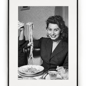May include: A black and white framed photograph featuring a person with wavy hair wearing a pinstriped blazer. The person is smiling while holding a fork with a long strand of spaghetti above a plate of pasta, with two glasses on the table.