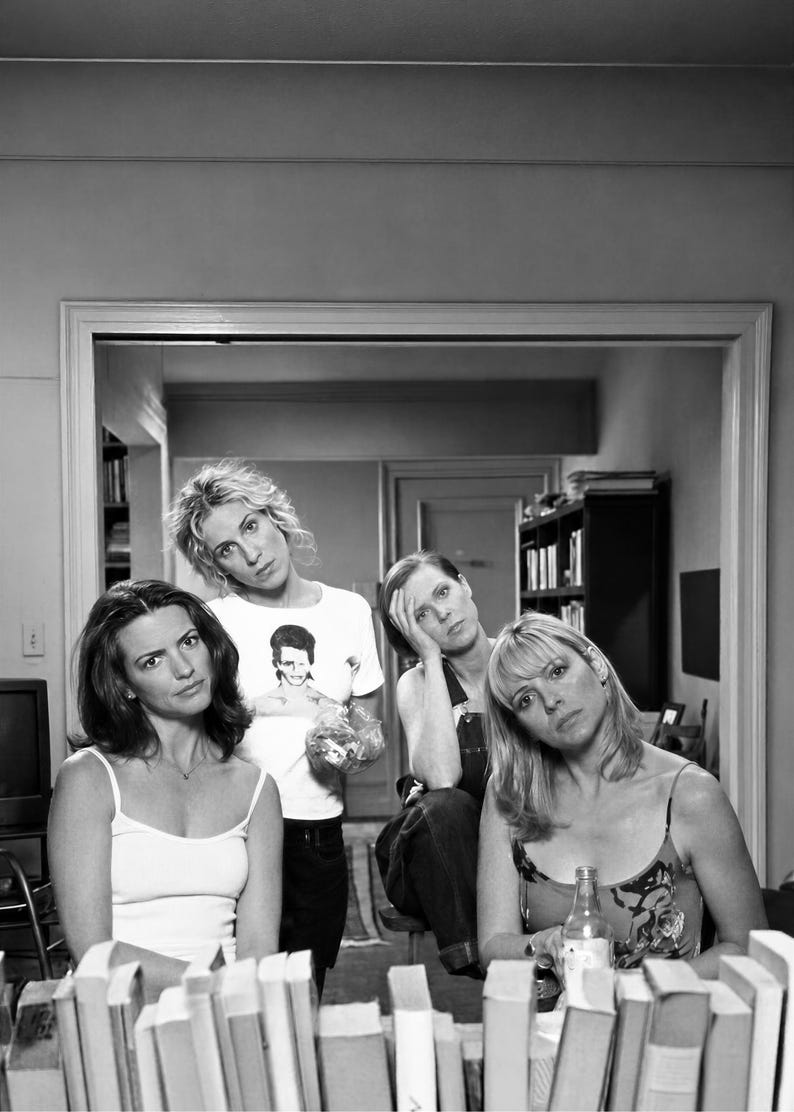 Pu&ograve; includere: Una fotografia in bianco e nero che ritrae quattro persone posizionate dietro una fila di libri. Una persona indossa una t-shirt bianca con una stampa grafica, un'altra una canotta bianca, una indossa una salopette e la quarta un top a fantasia.