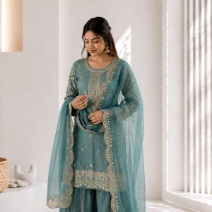 May include: A teal-colored embroidered salwar kameez set with a matching dupatta. The outfit features intricate gold embroidery along the neckline, sleeves, and hemline. The Shoplix logo is visible in the upper right corner.