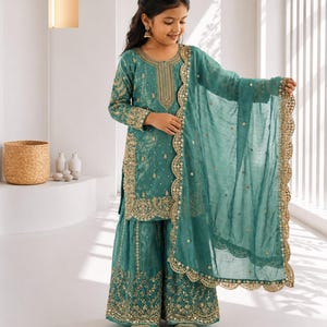 Könnte beinhalten: Ein teal-farbener, bestickter Sharara-Anzug mit goldenen Akzenten. Das Outfit umfasst eine langärmlige Tunika, eine Hose mit weitem Bein und einen passenden Dupatta. Der Anzug hat aufwendige goldene Stickereien am Ausschnitt, an den Ärmeln und am Hosenbund. Das Shoplix-Logo ist sichtbar.