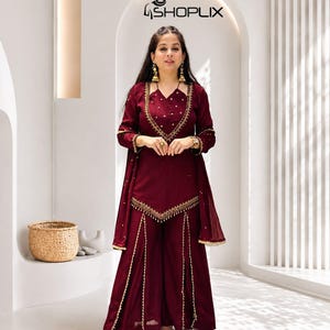 May include: A deep maroon three-piece outfit featuring a long-sleeved tunic with a V-neckline, gold embroidery, and a matching dupatta. The wide-leg trousers have vertical gold trim detailing. The text SHOPLIX is visible at the top of the image.
