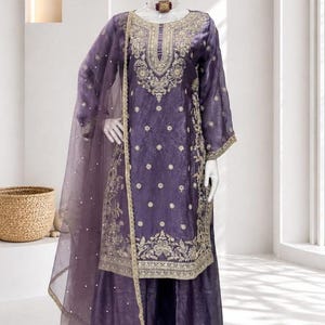 Peut inclure: Ensemble salwar kameez violet brodé avec un dupatta assorti. La tenue présente des broderies dorées complexes sur l'encolure, les manches et l'ourlet. Le mannequin porte un couvre-chef doré et un collier. Le logo Shoplix est visible.