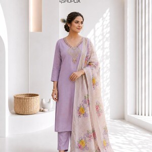 Pode incluir: Um conjunto salwar kameez lilás com decote em V, detalhes bordados e calças a condizer. O conjunto inclui um lenço com estampa floral. A imagem mostra uma mulher usando o traje.