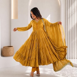 May include: Mustard yellow tiered dress with long sleeves and a matching scarf. The dress has a V-neck and a flowing, flared skirt. The outfit is paired with matching trousers and embellished heels. The Shoplix logo is visible in the upper right corner.