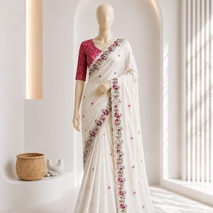 May include: A cream-colored saree featuring a dark pink blouse with a textured pattern. The saree is adorned with a floral border in shades of pink and green, with matching floral motifs scattered across the fabric. The text SHOPLIX is visible.