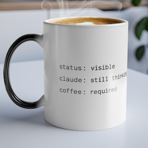 Puede incluir: Taza de café de cerámica blanca con asa negra, llena de café y vapor que se eleva desde la parte superior. La taza tiene el texto "status: visible, claude: still thinking, coffee: required" impreso en el lateral.