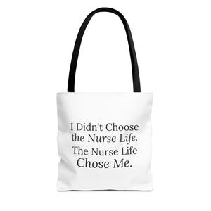 Op de afbeelding: Een witte tote bag met zwarte hengsels voorzien van de zwarte tekst: I Didn't Choose the Nurse Life. The Nurse Life Chose Me. De tas heeft een rechthoekige vorm met een platte bodem en twee stevige schouderbanden aan de bovenzijde.