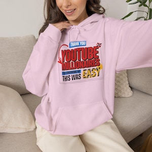 May include: A light pink hooded sweatshirt featuring a graphic print with the text "THANK YOU YOUTUBE MILLIONAIRES FOR CONVINCING ME THIS WAS EASY". The design includes red, blue, black, and yellow lettering with a red arrow and a crossed-out circle.