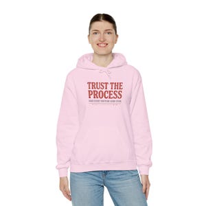 May include: A light pink hooded sweatshirt featuring the text "TRUST THE PROCESS" in bold red capital letters, followed by the smaller text "SAID EVERY YOUTUBE GURU EVER" in a dark font, with a front kangaroo pocket and long sleeves.