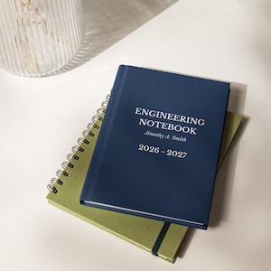 May include: A dark blue hardcover notebook with the white text ENGINEERING NOTEBOOK, Jimothy A. Smith, and 2026-2027 printed on the front cover, resting on top of a green spiral-bound notebook with a black elastic band closure.
