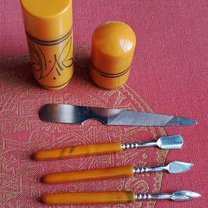 Może przedstawiać: Zestaw do manicure składający się z dwóch cylindrycznych pojemników w kolorze musztardowym z czarnymi liniami, metalowego pilnika do paznokci oraz trzech metalowych narzędzi do pielęgnacji z pasującymi musztardowymi uchwytami, w tym kopytka.