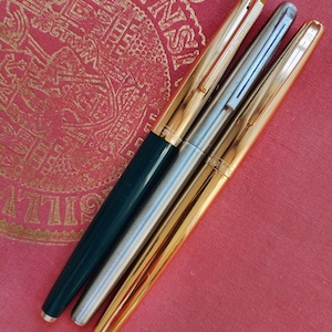 Könnte beinhalten: Drei Füllfederhalter, die diagonal angeordnet sind. Der linke Stift hat einen dunkelgrünen Schaft und eine goldene Kappe. Der mittlere Stift ist aus gebürstetem Silber mit passender Kappe und Clip. Der rechte Stift ist komplett goldfarben.