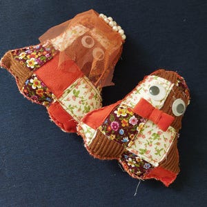 May include: Two stuffed fabric figures featuring patchwork designs in floral patterns, solid orange, and brown corduroy. One figure wears an orange mesh veil with white beads, while the other features two googly eyes and a small orange fabric bow tie.