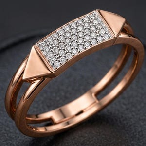 May include: Rose gold ring with a rectangular center encrusted with small, sparkling diamonds. The ring features a geometric design with a double band and triangular accents on each side of the diamond-studded rectangle. The ring is set against a dark background.