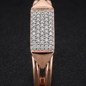 May include: Rose gold ring with a rectangular center filled with small, sparkling white stones. The ring has a geometric design with triangular accents at the top and bottom of the rectangular center. The band is split, creating an open design.