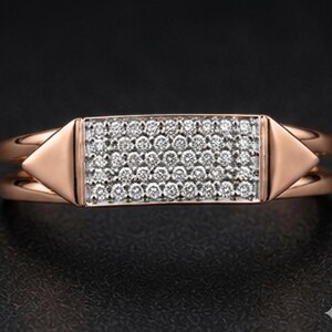 May include: Rose gold ring with a rectangular center encrusted with small, sparkling white gemstones. The ring features a double band design and triangular accents on each side of the gemstone setting.