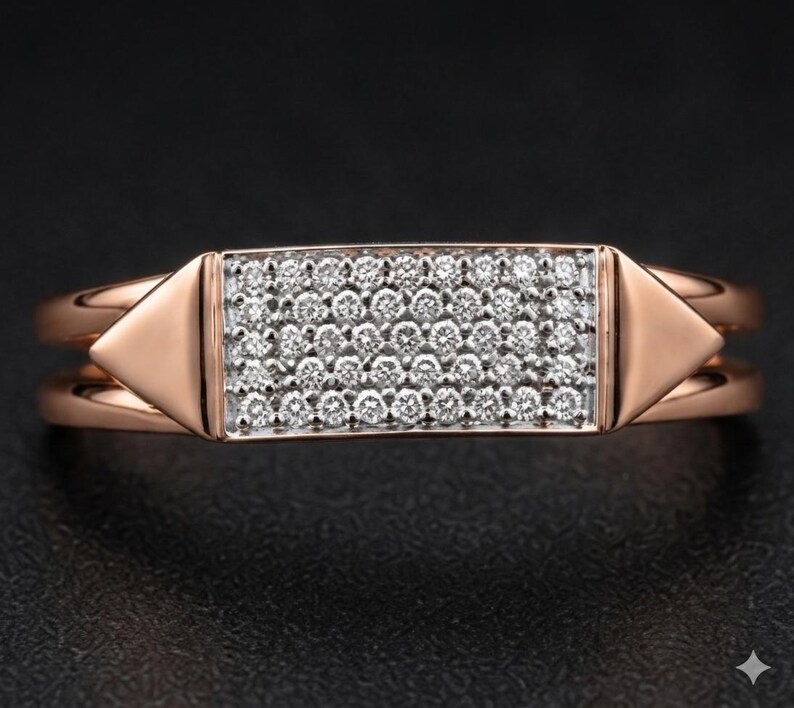 May include: Rose gold ring with a rectangular center encrusted with small, sparkling white gemstones. The ring features a double band design and triangular accents on each side of the gemstone setting.