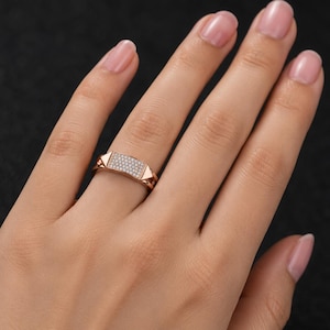 May include: Rose gold ring with a rectangular center encrusted with small, clear gemstones. The ring features geometric, triangular accents on each side of the gemstone panel. The ring is shown on a person's finger.