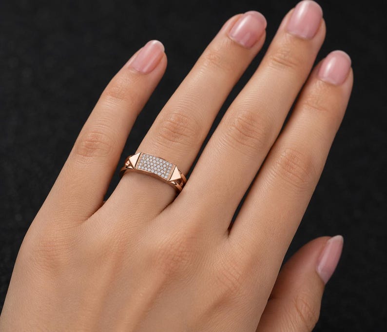 May include: Rose gold ring with a rectangular center encrusted with small, clear gemstones. The ring features geometric, triangular accents on each side of the gemstone panel. The ring is shown on a person's finger.