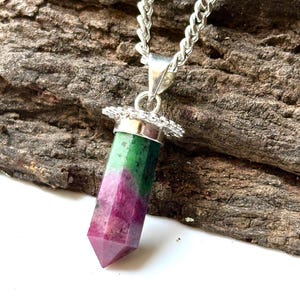 May include: A pendant necklace featuring a hexagonal, pointed ruby and zoisite crystal stone with a vibrant magenta and deep green color palette. The stone is secured by a silver-toned metal cap with a decorative, scalloped edge and hangs from a silver-toned chain.